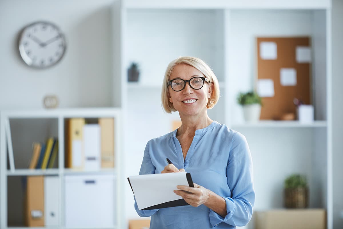 Blond woman standing in the office with a clock on the background.