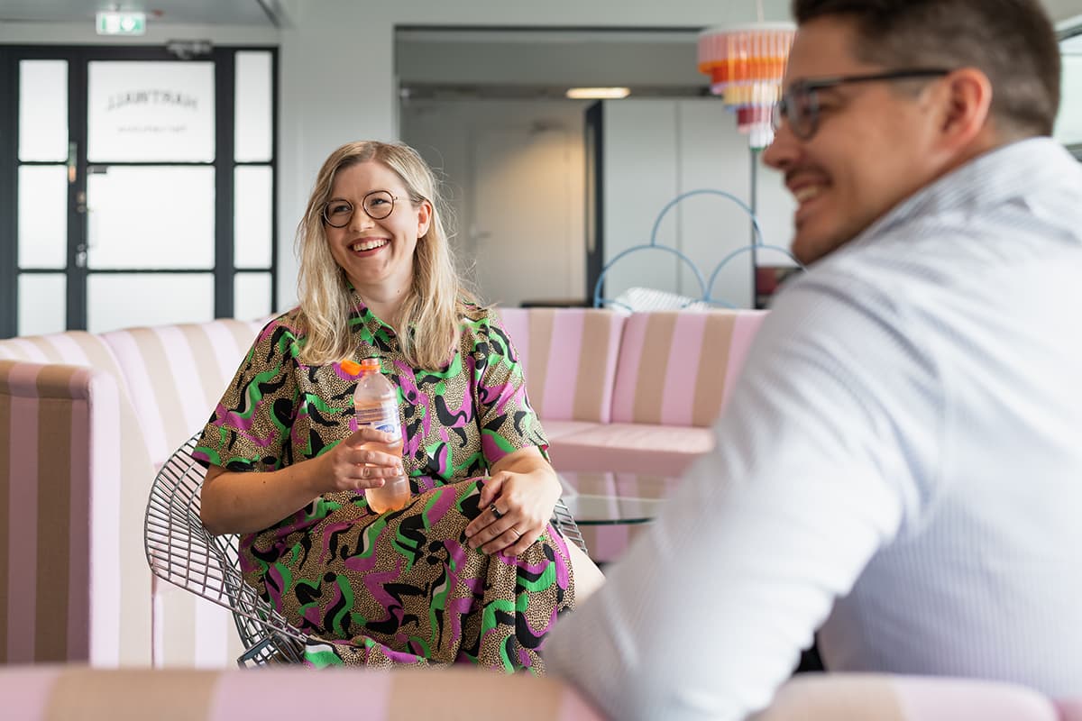 Nainen istuu sohvalla kädessään Hartwallin virvoitusjuomapullo, etualalla mies istumassa sivuttain.